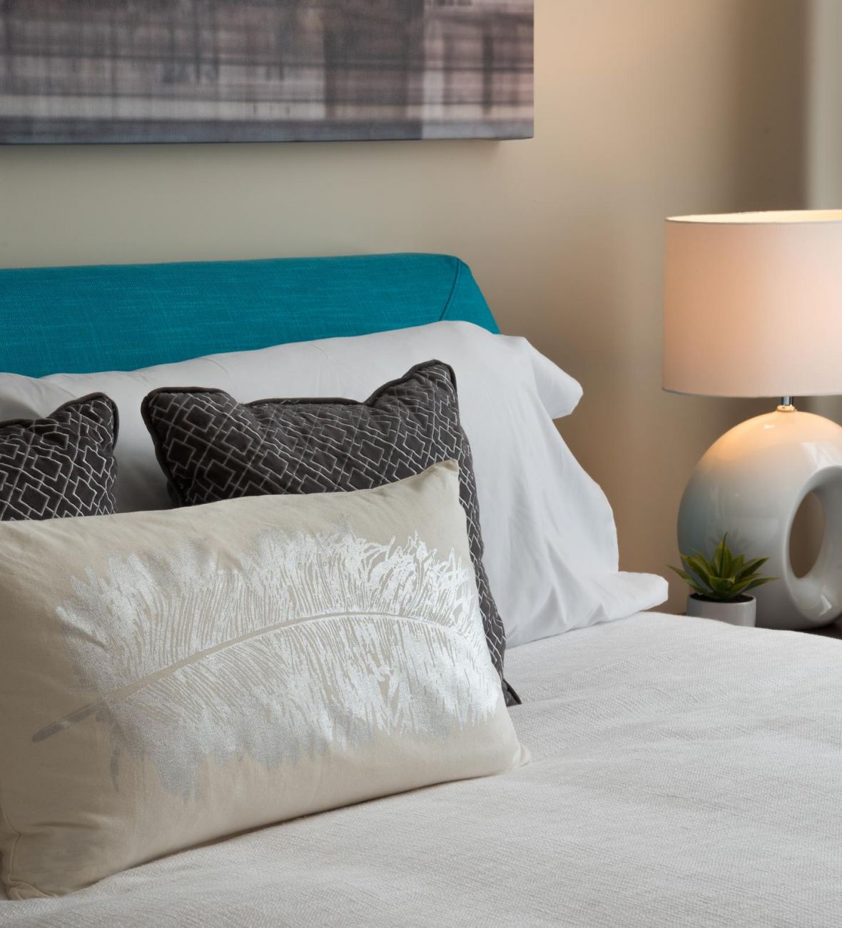 Pinnacle Apartments Modern bedroom with teal headboard, decorative pillows, and a nightstand with a lamp, clock, and small plant.