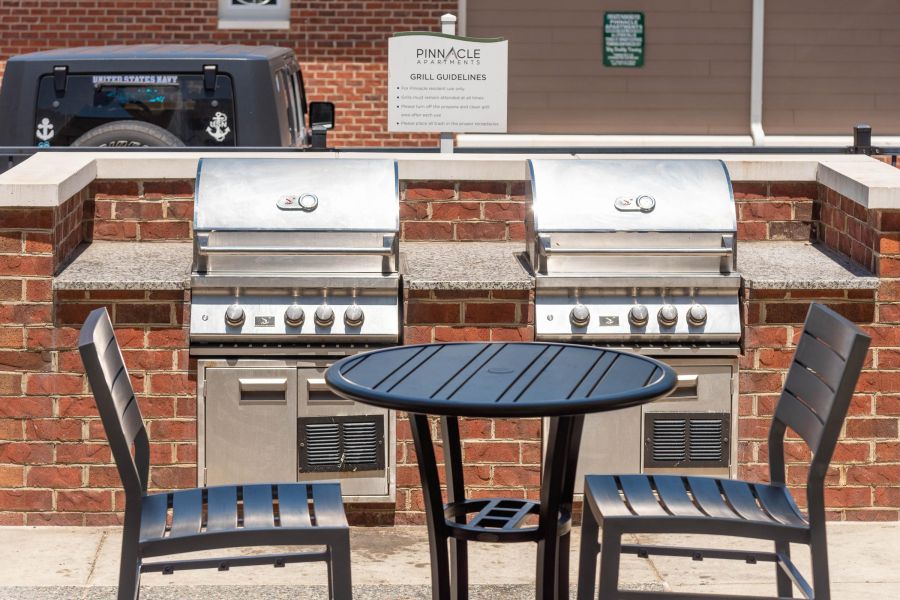 Pinnacle Apartments Two stainless steel grills behind a round black table with two chairs on a patio near a brick wall.
