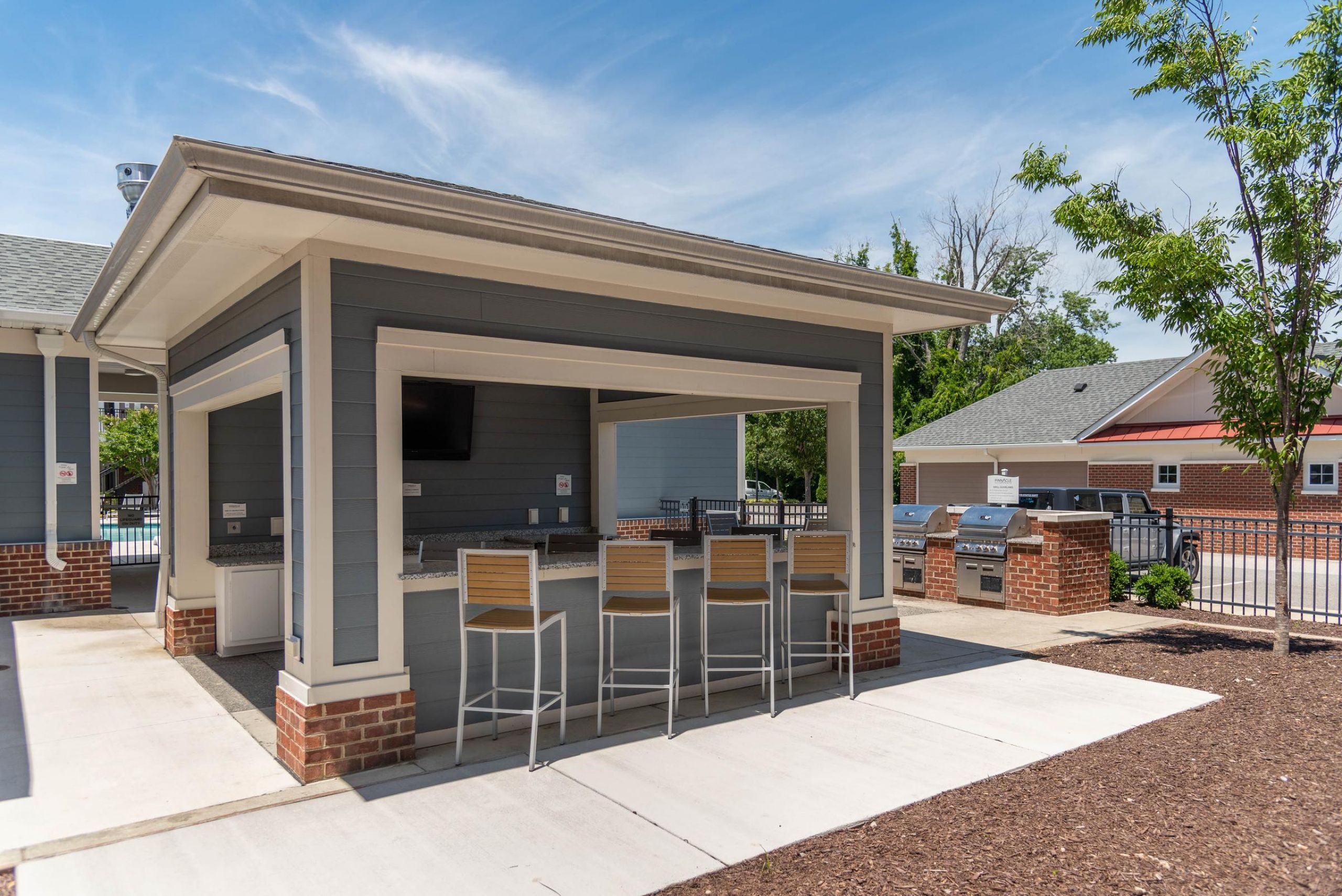 Pinnacle Outdoor covered bar area with stools, two grills, and a TV, next to a pool and a tree on a sunny day.