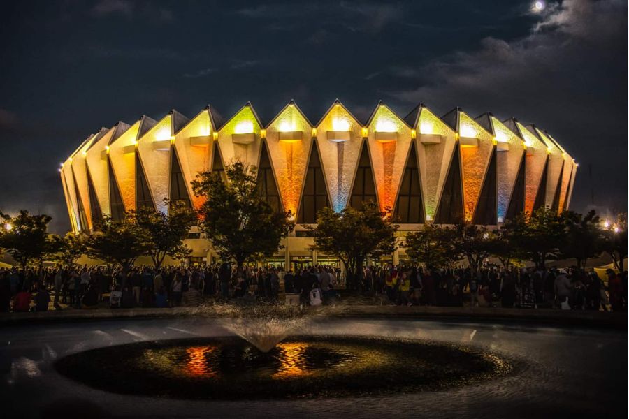 Pinnacle Apartments A brightly lit arena with a unique roof and many people gathered outside at night, with a fountain in front.