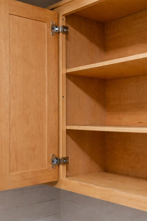 Pinnacle Apartments Open wooden kitchen cabinet with empty shelves and metal hinges on the door, against a tiled wall.