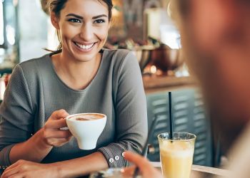 Pinnacle Apartments Smiling woman holding a cup of coffee, sitting at a table with another person in a café.