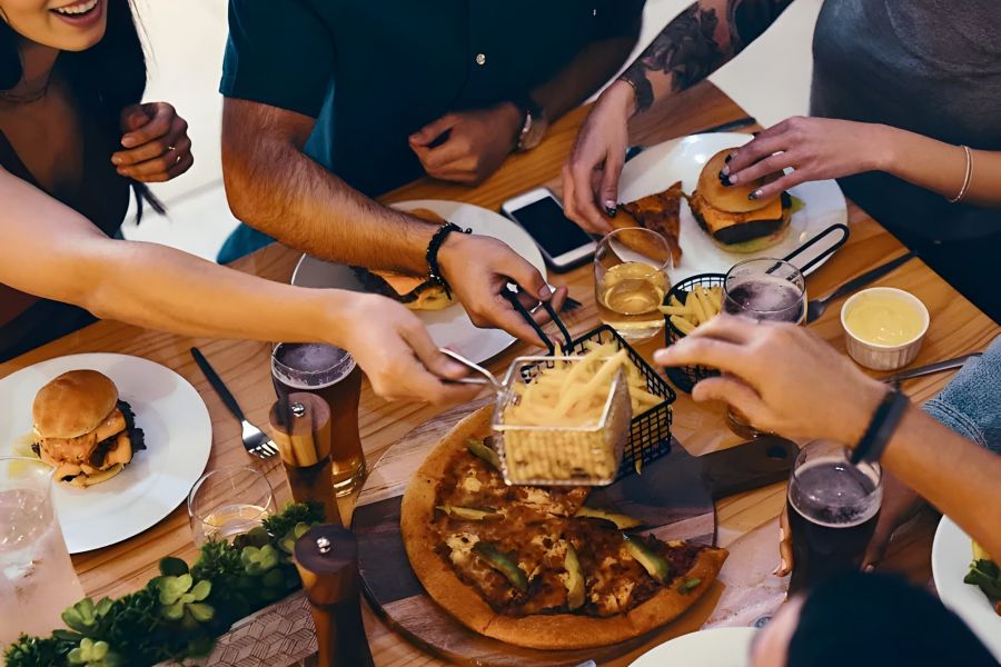 Pinnacle Apartments Friends share pizza, burgers, and fries at a wooden table, enjoying a meal and drinks together.