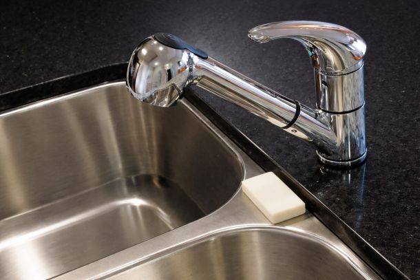 Pinnacle Apartment Chrome kitchen faucet over a double stainless steel sink with a bar of soap on the black countertop edge.