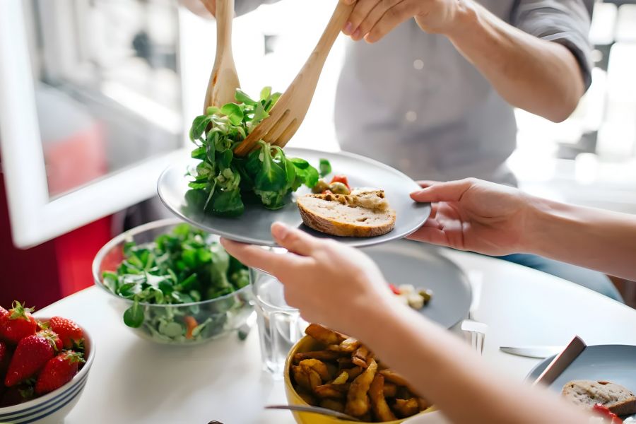 Pinnacle Apartments Person serving salad onto a plate at a table with bread, strawberries, and other dishes.