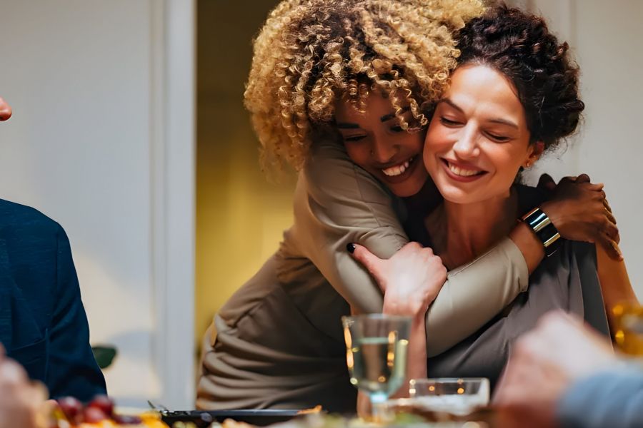 Pinnacle Apartments Two women happily hug each other at a dinner table, smiling with food and drinks in front of them.