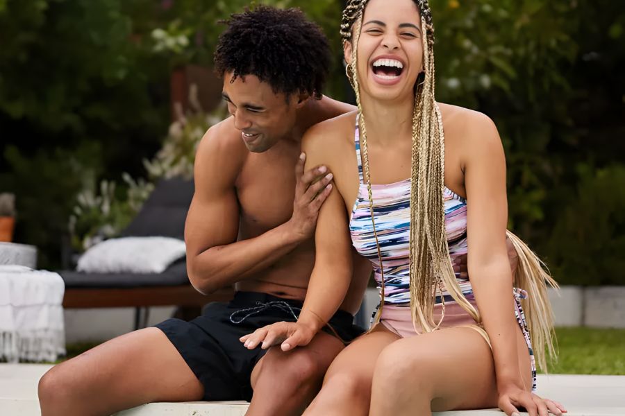 Pinnacle Apartments A smiling couple sits on the edge of a pool, laughing and enjoying a sunny day outdoors.