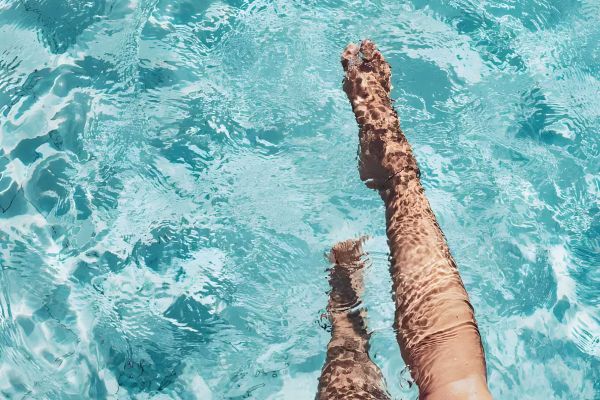 Pinnacle Apartments Two legs are partially submerged in a clear blue swimming pool with sunlight reflecting on the water's surface.